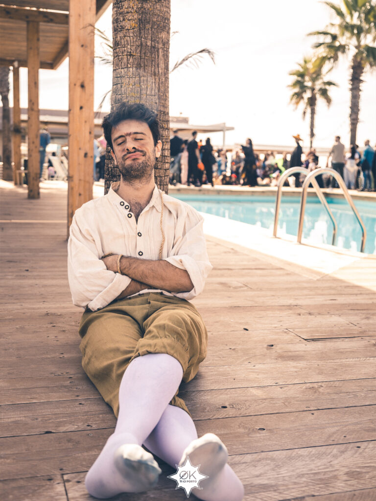 Un cattivo in camicia color crema e pantaloni chiari, appoggiato a una palma con le braccia incrociate